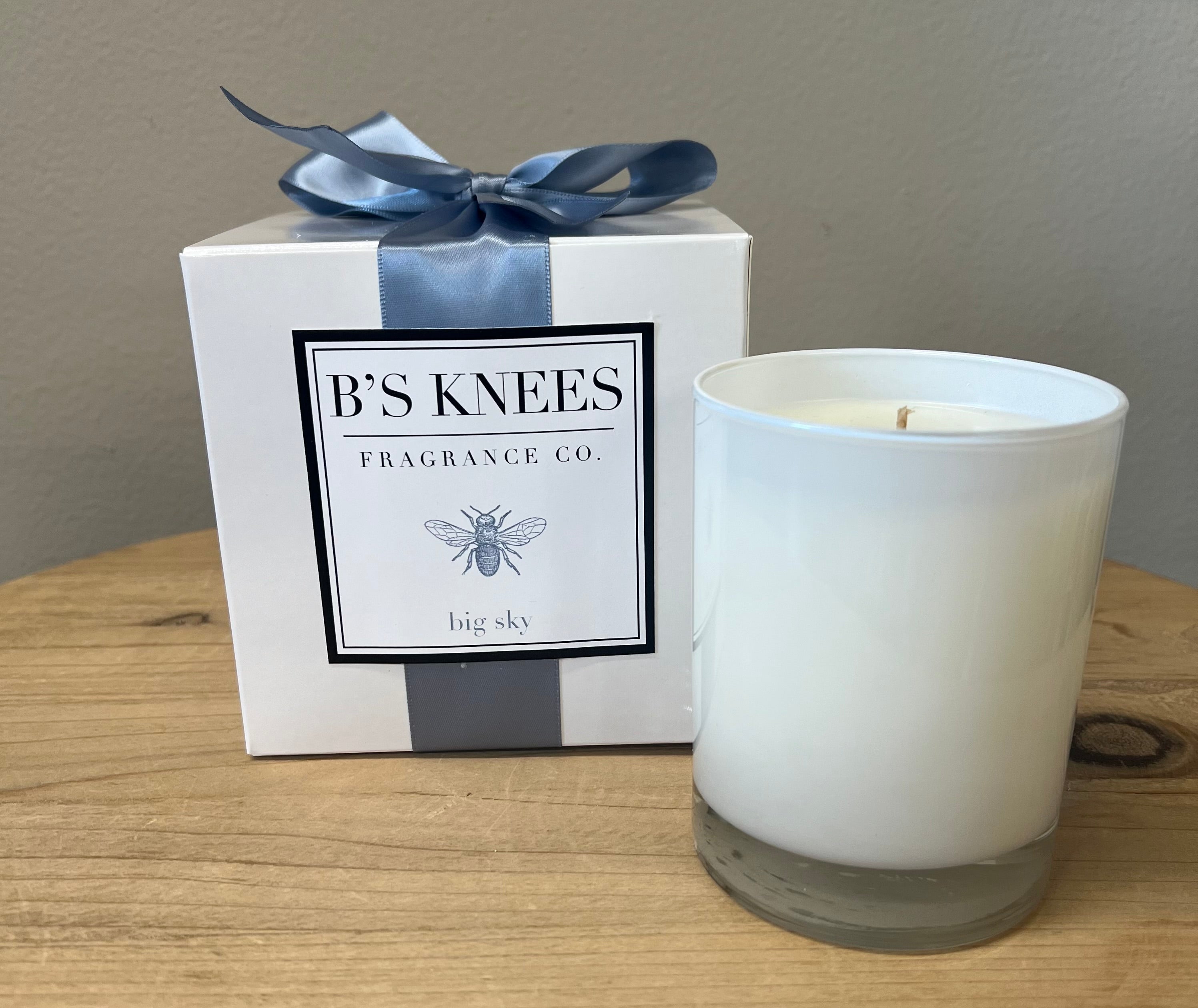 A clear glass candle with a white label that reads 'B's Knees FRAGRANCE COMPANY Big Sky', presented in a white box with a blue satin ribbon and a blue bow on top.