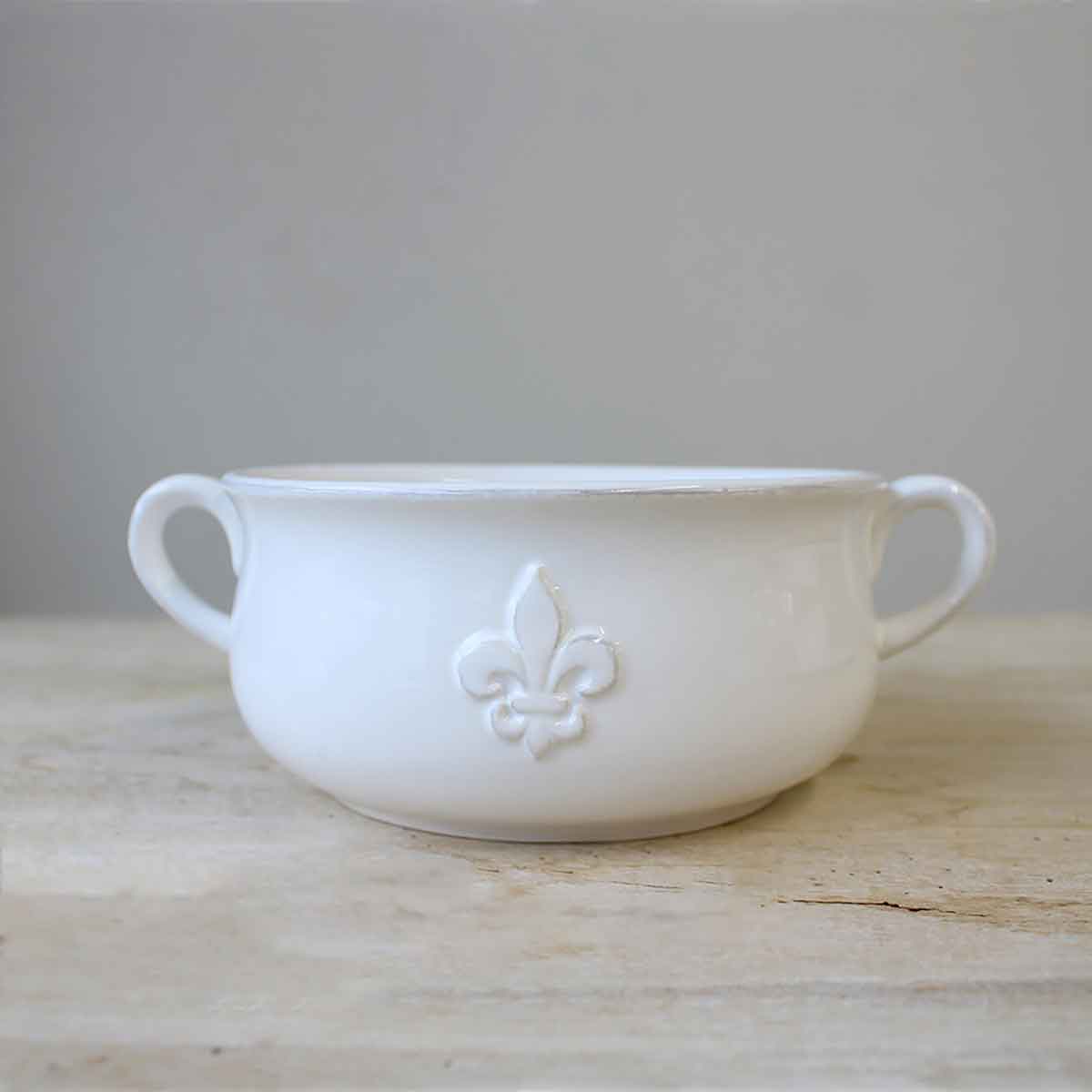 A white ceramic gumbo bowl with double handles and a Fleur de Lis embossing on the side