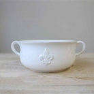 A white ceramic gumbo bowl with double handles and a Fleur de Lis embossing on the side